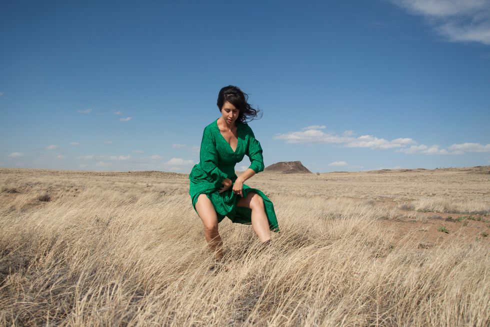 Miracle, wearing a forest green dress, dances in tall grass, a vast landscape behind her