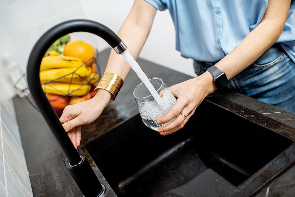 A woman fills up a glass cup with water at a sleek black sink. A bowl of fruit sits beside the sink.
