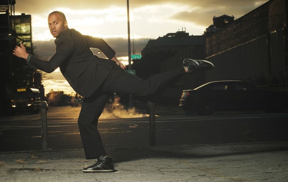 White on a city street, in an exaggerated running positionu2014one leg back in attitude and same arm extended in front of him. He looks at the camera, and wears a black suit.