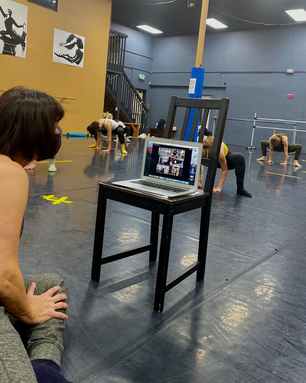 A group of students stretch, while Cooper sits on the floor looking at a Zoom screen full of students with her computer propped up on a chair