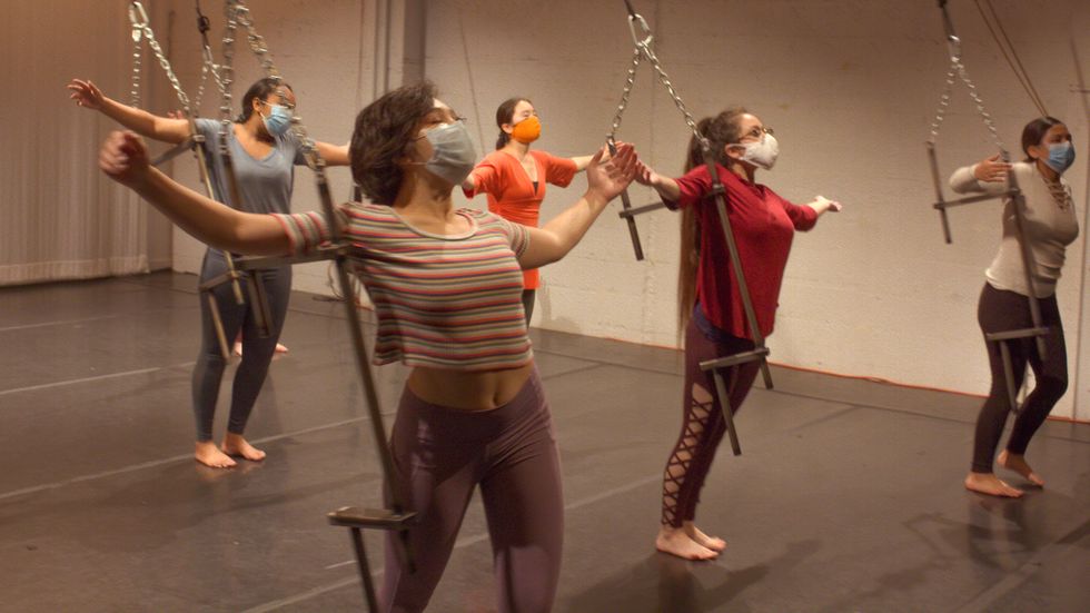 Five masked teen girls hang on contraptions hanging from the ceiling in a studio