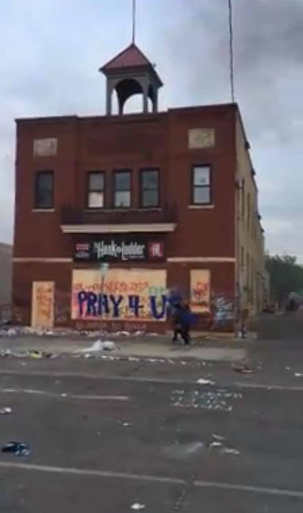 A blurry image of a boarded up building with a sign that says "Pray 4 U." The street is littered with debris.