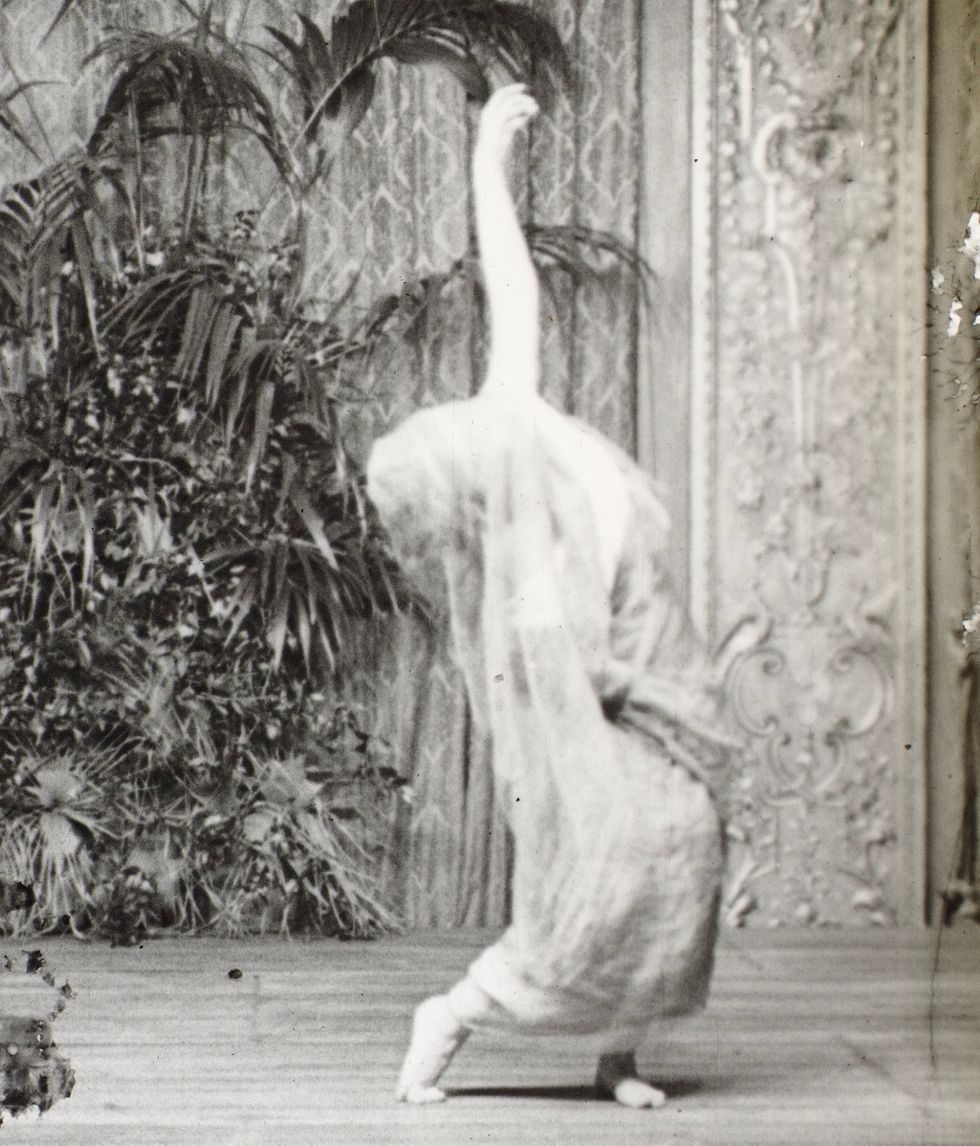A grainy black and white images of Noyes, in a white tunic draped over her head, leaning to the side.