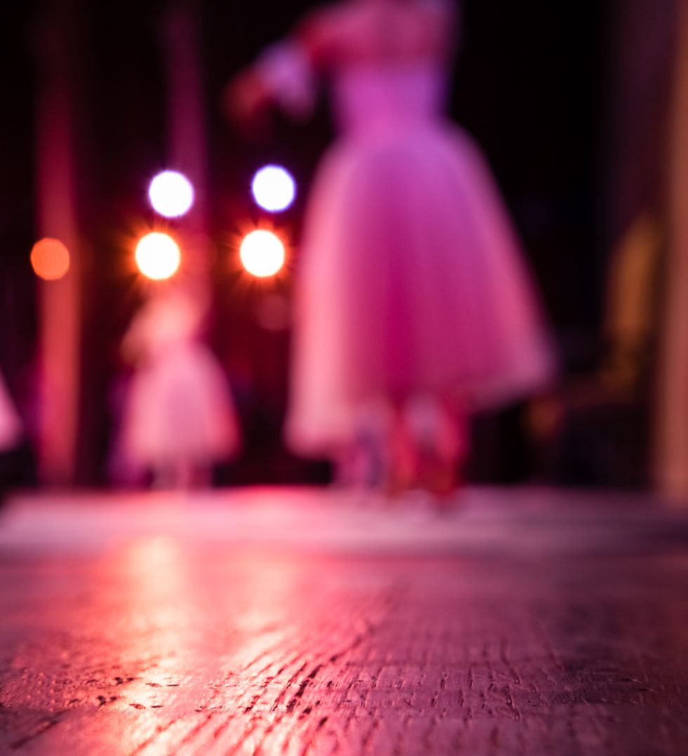 Blurred ballerinas in long white tutus dance on a wooden floor and are viewed from backstage.