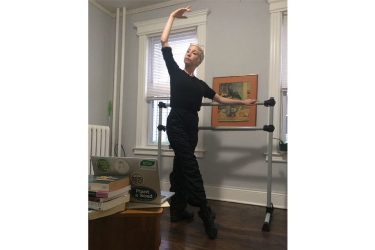 "Rosner, wearing all black, stands at a small barre in her home, demonstrating a tendu front to her laptop screen.