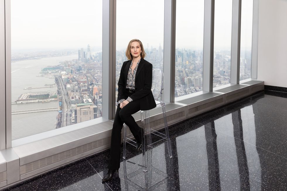 Whelan, in business casual wear, sits on a clear chair in a window-filled room overlooking the New York City skyline