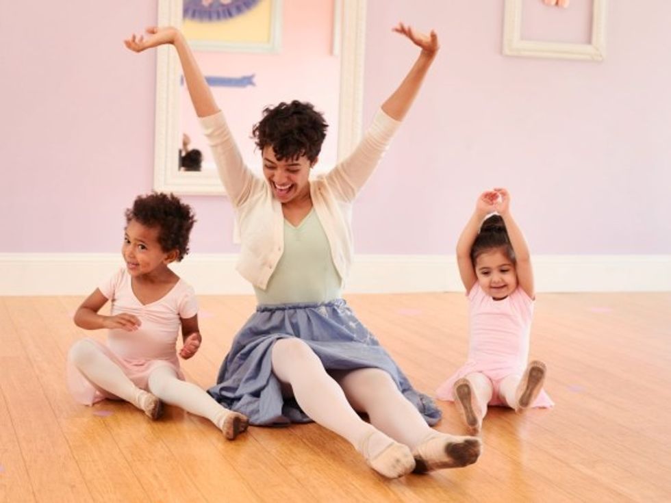 A teacher sits on the floor, arms raised to the sky in delight. She's flanked by two toddler-aged girls who copy her.