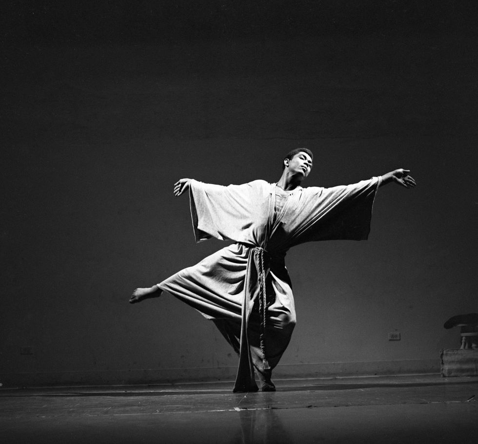 A black-and-white image of Alvin Ailey onstage. He is wearing a loose robe and is in a low arabesque in pliu00e9 with his arms extended sideways