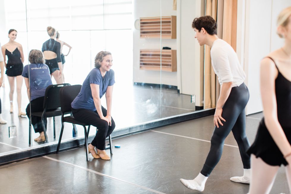 Ringer, smiling in a blue shirt and black pants, sits in a chair at the front of the studio, smiling at the teen ballet students in front of her