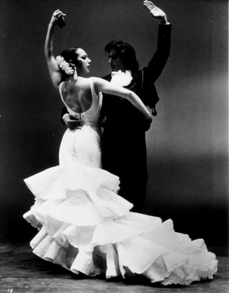 A black and white photo of a male and a female Flamenco dance pair. They dance together, wrapping their arms around each other's waists.