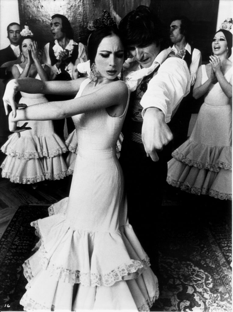 A black and white photo of a male and a female Flamenco dance pair. They dance together in a crowded room.