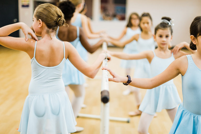 Young girls in blue leotards and skirts hold either side of a barre, with their outside hand on their shoulder. They are mid-plié.