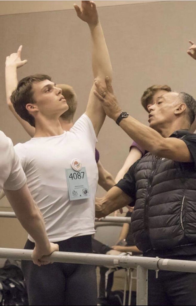 Olivier Muñoz holds the left arm of a male ballet student at the barre as he lifts it in fifth position. The boy wears a white T-shirt and black tights, while Muñoz wears a black puffer vest and black pants.