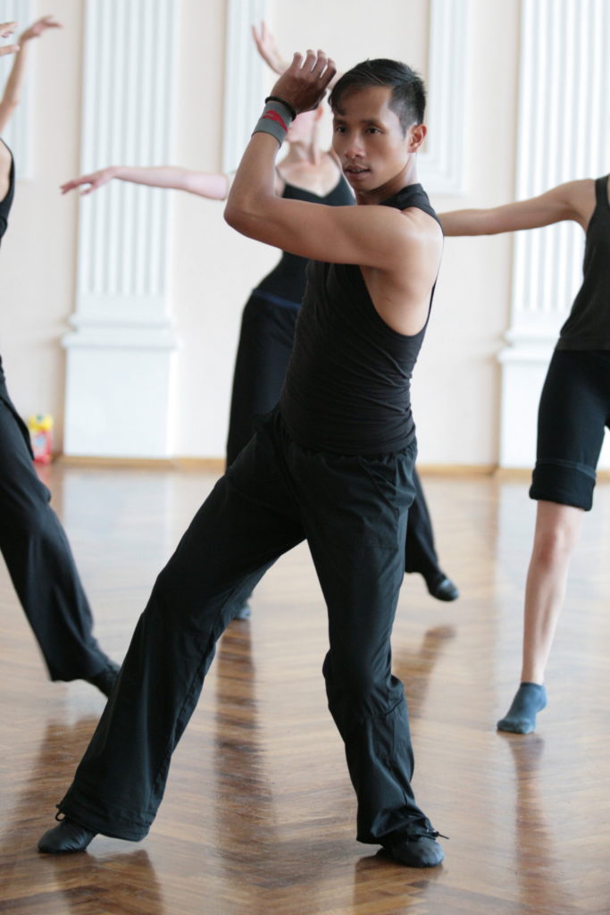 Eddy Ocampo wears black yoga pants, a black tank top and jazz shoes leads a group of students through a jazz class. He twists to the right, swinging his left arm across his body, and pliés deeply on both legs.