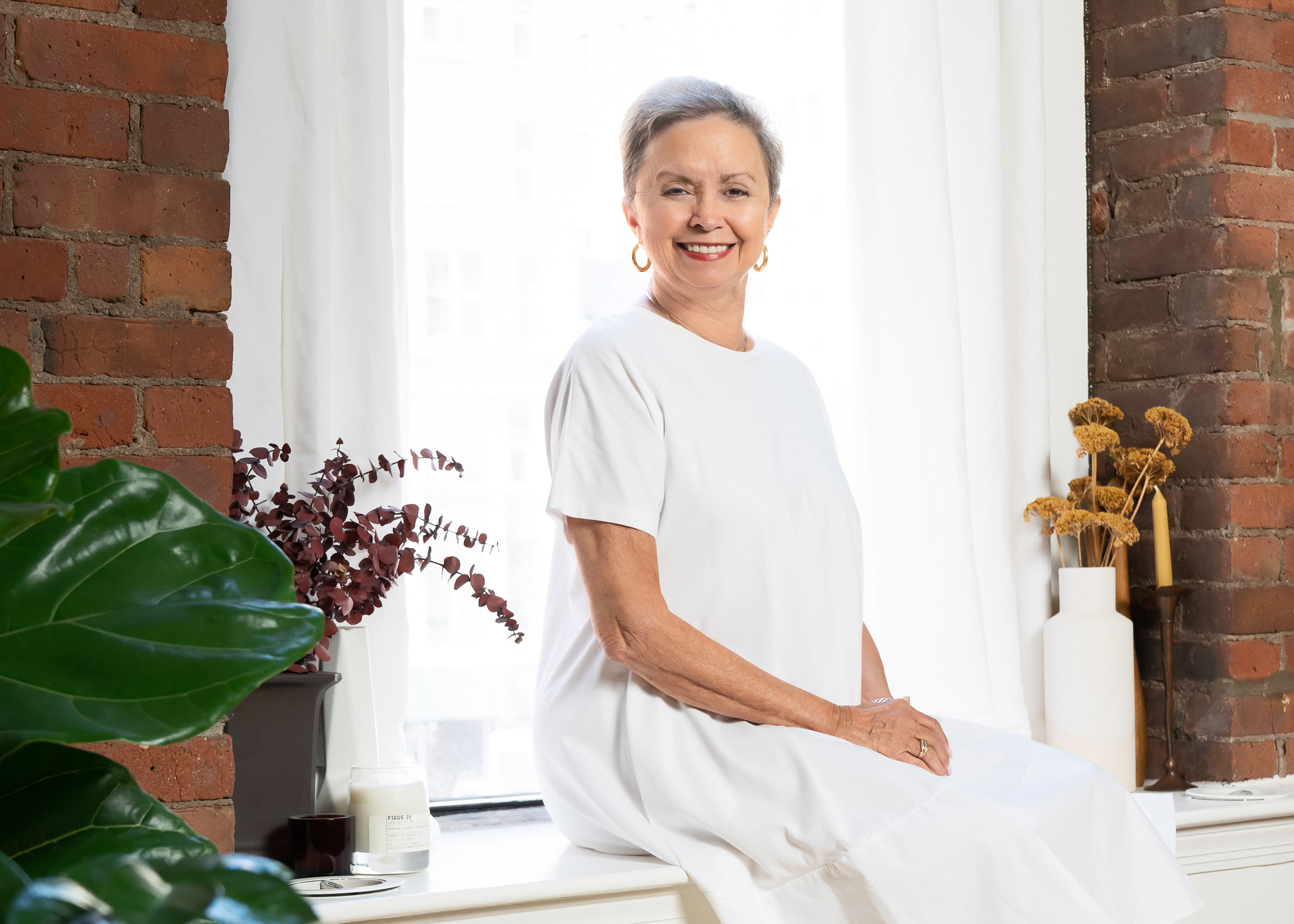 Kay Mazzo sits on a window sill and smiles toward the camera. She wears a white dress and folds her hands in her lap.