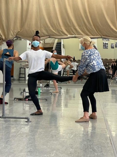 During a ballet class, Kristine Elliot stands next to a male ballet student and holds his left lower leg as he extends it into a 90 degree développé. She wears a blue and white floral top, black tights, a black ballet skirt and tan jazz shoes. The student wears a white T-shirt, black tights and brown ballet slippers.