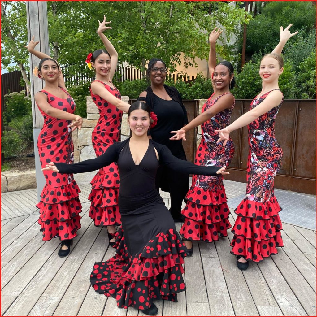 Yvonne Gutierrez, in all black, poses in the back with her Spanish dancers. The young female dancers are dressed in bright red and black performance dresses, adorned with floral patterns and polka dots. Their hair is pulled back into sleek low buns with adorning flowers. The group poses outside on a wooden patio in front of trees and bushes.