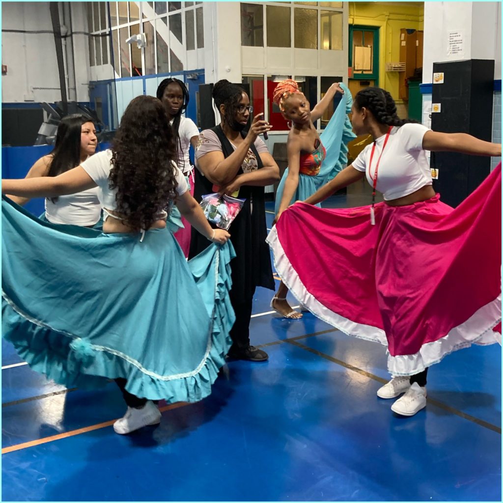 Yvonne Gutierrez stands in the middle of a circle formed by her Pathways2Graduation students. The dancers, all young adult females, wear white or floral print tops and long blue or pink skirts, poses around Yvonne. They dance in a gymnasium
