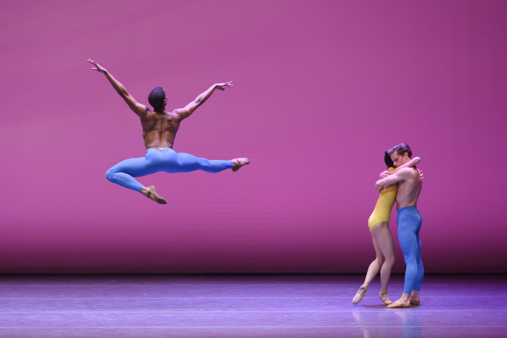 A ballet performance scene on a magenta-lit stage. A male dancer is photographed midair in a stag position from behind, his arms in a V. To his right, a man and woman embrace each other.