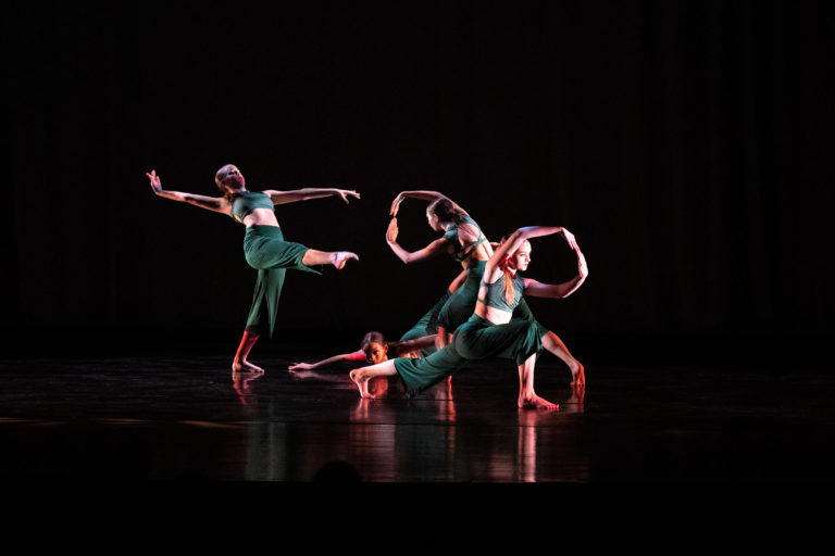 Four young women in green pants and crops tops form a cluster of shapes in a modern dance performance.