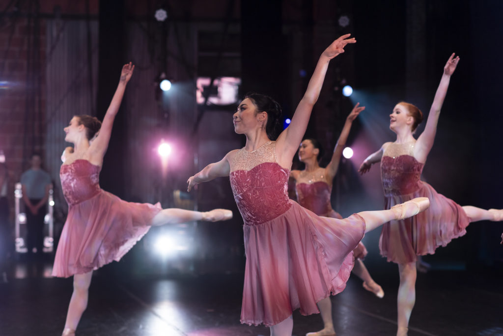 Four ballet dancers in rose-colored contemporary ballet dresses in a low piqué arabesque position.