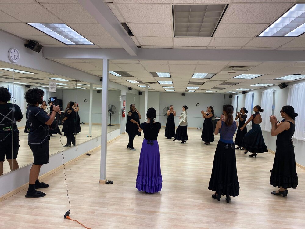In a light gray dance studio with light wood flooring, Yvonne Gutierrez leads a class of all-female students in a traditional Spanish dance class. Yvonne is dressed in all black with flamenco heels, and the students wear mostly black in leotards, flamenco heels, and long flamenco skirts, their hair pulled back into braids, buns, or ponytails. A cameraman stands to the left of the photo toward the mirror, recording the session.
