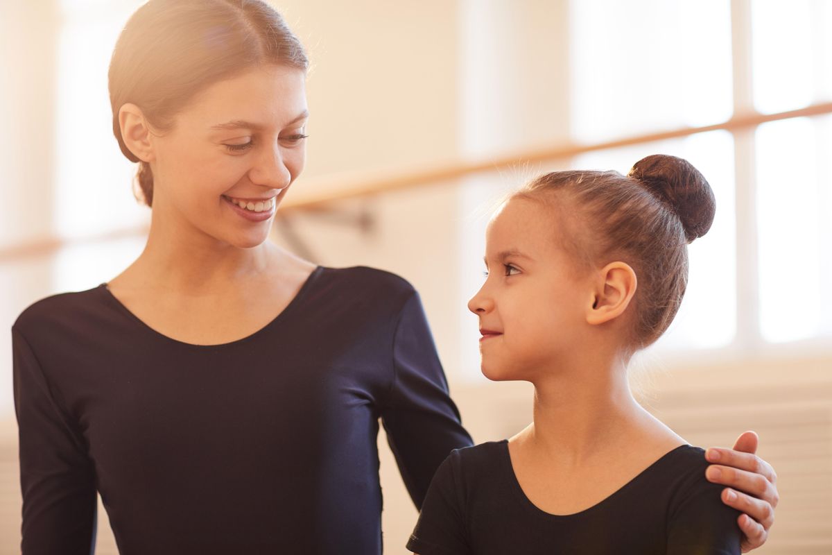 A ballet teacher in he early twenties wraps her left arm around a young ballet student and looks down at her with a caring smile, which the child returns. They both wear black leotards and sit in a sunny ballet studio.