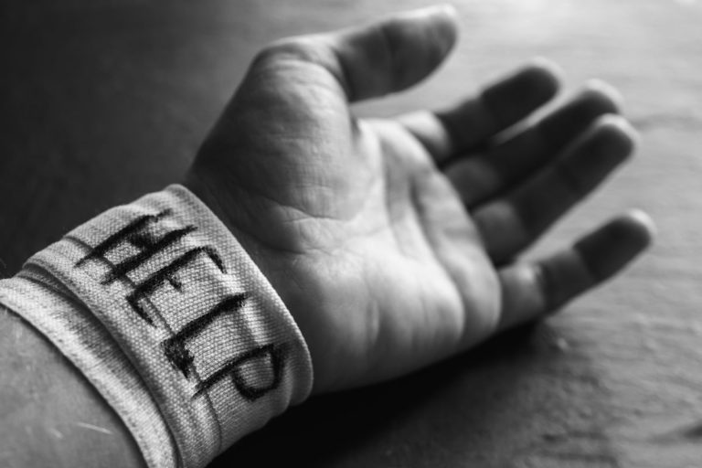 A black and white photo of a wrist, lying on a table, that has a bandage around it saying 'help'