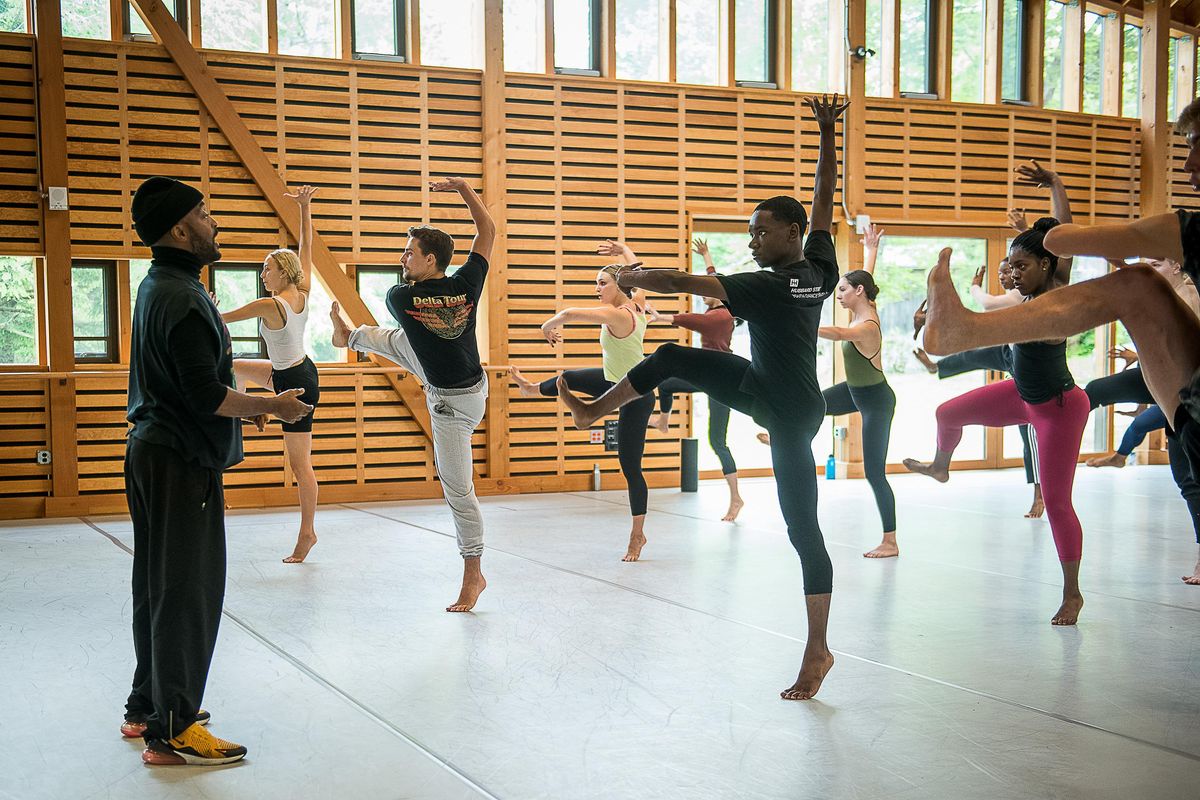 Darrell Grand Moultrie, a young Black man wearing yellow sneakers, black sweats and turtleneck and a black beanie, teaches a contemporary class, with young adult dancers balancing on one leg, other leg bent and to the side. They push one arm up to the ceiling, the other forward. They are in a light filled barn studio.
