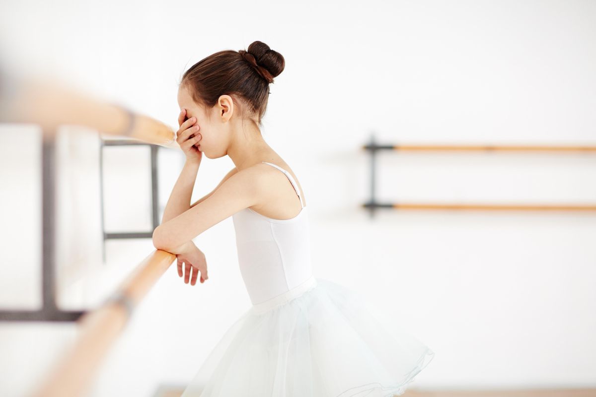 A young girl in a white leotard and tutu leans against a ballet barre with her head in her hand.