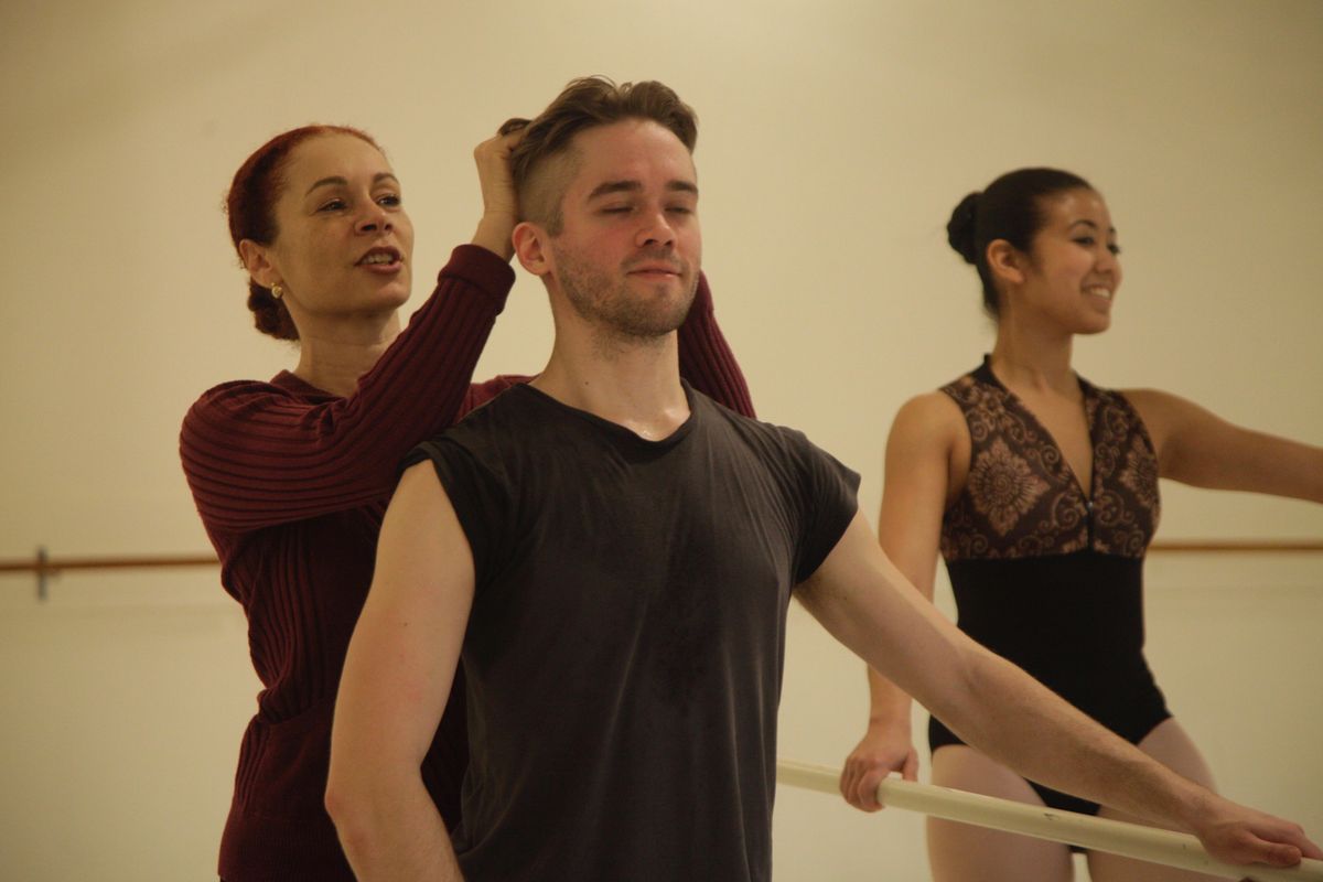 Carole Alexis stands behind a young adult man at the barre, adjusting his head placement. She is a light-skinned Black woman with red hair, wearing a red sweater
