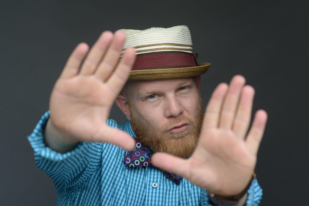 Rutledge, a middle-aged redheaded man with a beard, wears a blue checkered shirt, a colorful dotted bow tie and a hat. He reaches his hands towards the camera, framing his face