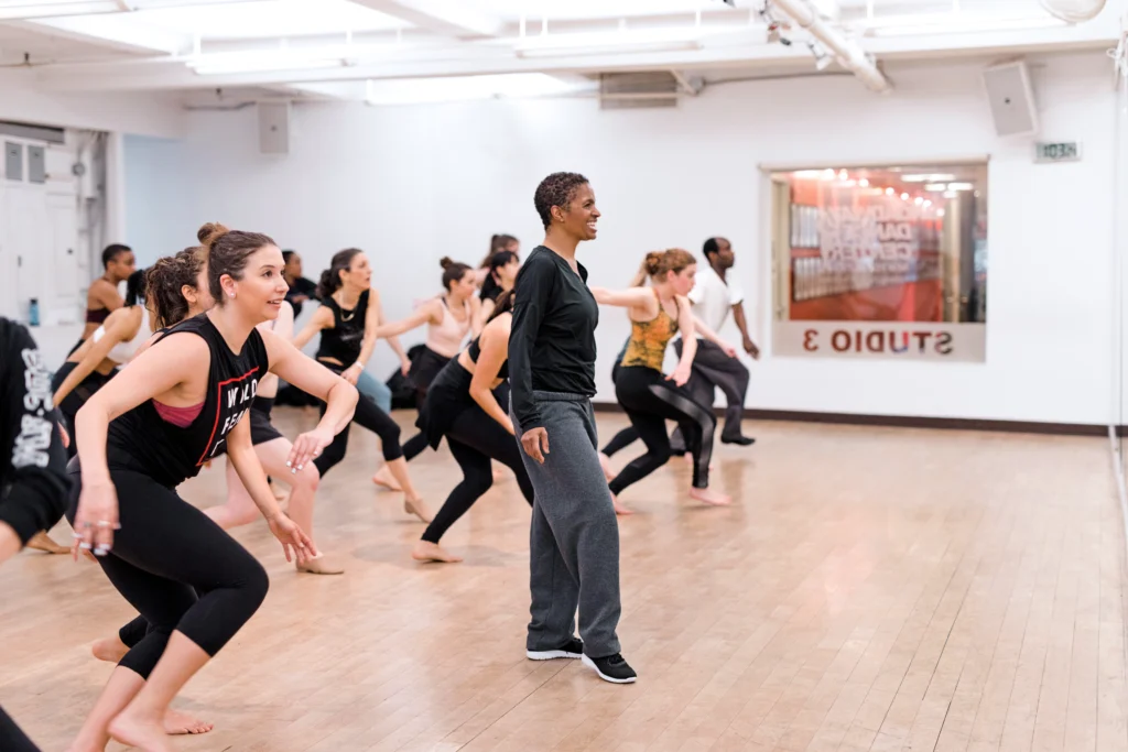 Sheila Barker, wearing grey leggings, a black long-sleeved top, and black sneakers, smiles as she teaches a class at Broadway Dance Center.