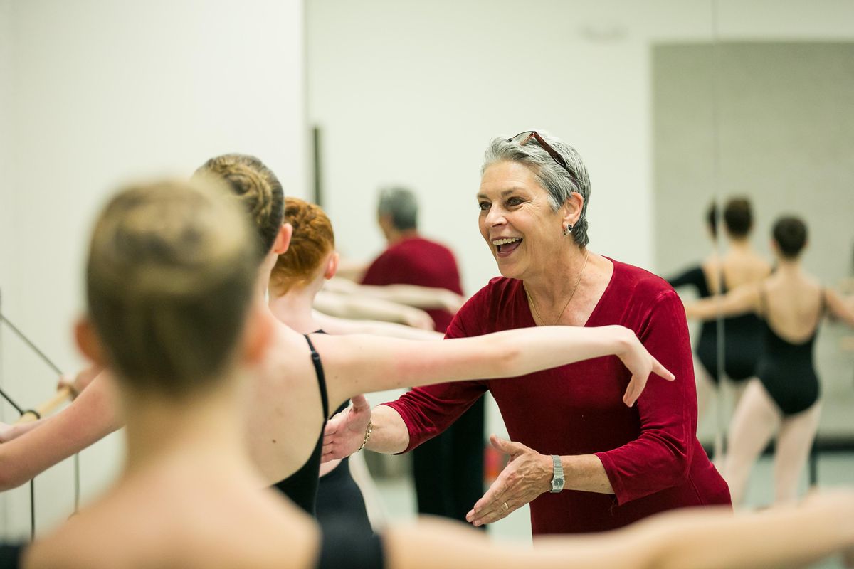 Bennett, wearing a red long-sleeved shirt and with her grey hair cut short, smiles and gestures at a young woman at the barre, who has her back to the camera and her arm in second position, along with her classmates.