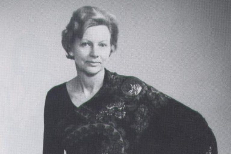 A middle aged Kai sits in a chair, posing with one hand on her hip. She wears a black patterned dress with fringes that hang off her arm.