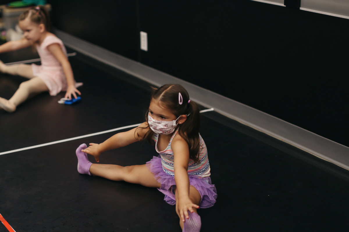 A toddler-aged girl sits in the side splits, reaching for her toes. She wears a patterned mask, a purple tutu and her hair in pigtails