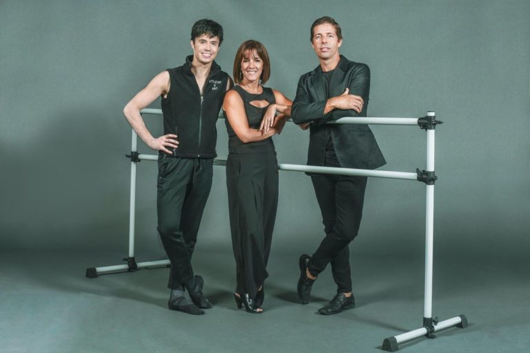 Joseph Gatti, Zoica Tovar and Andreas Estevez stand close together around a portable ballet barre in front of a gray backdrop. They are each wearing a black shirt, black pants and black shoes, and stand on one foot with their other foot crossed over, smiling towards the camera.