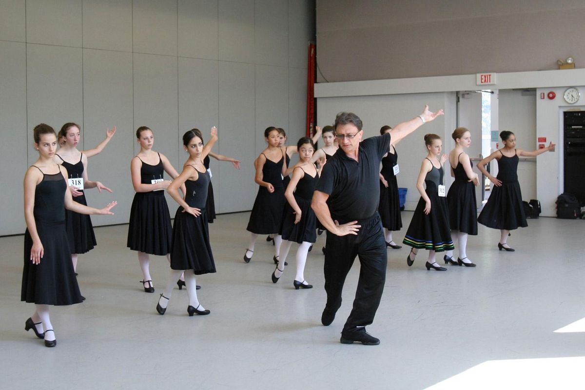 A man in all black demonstrates a plié tendu back with one arm out and the other on the hip to a group of teenage girls in black leotards and character skirts.