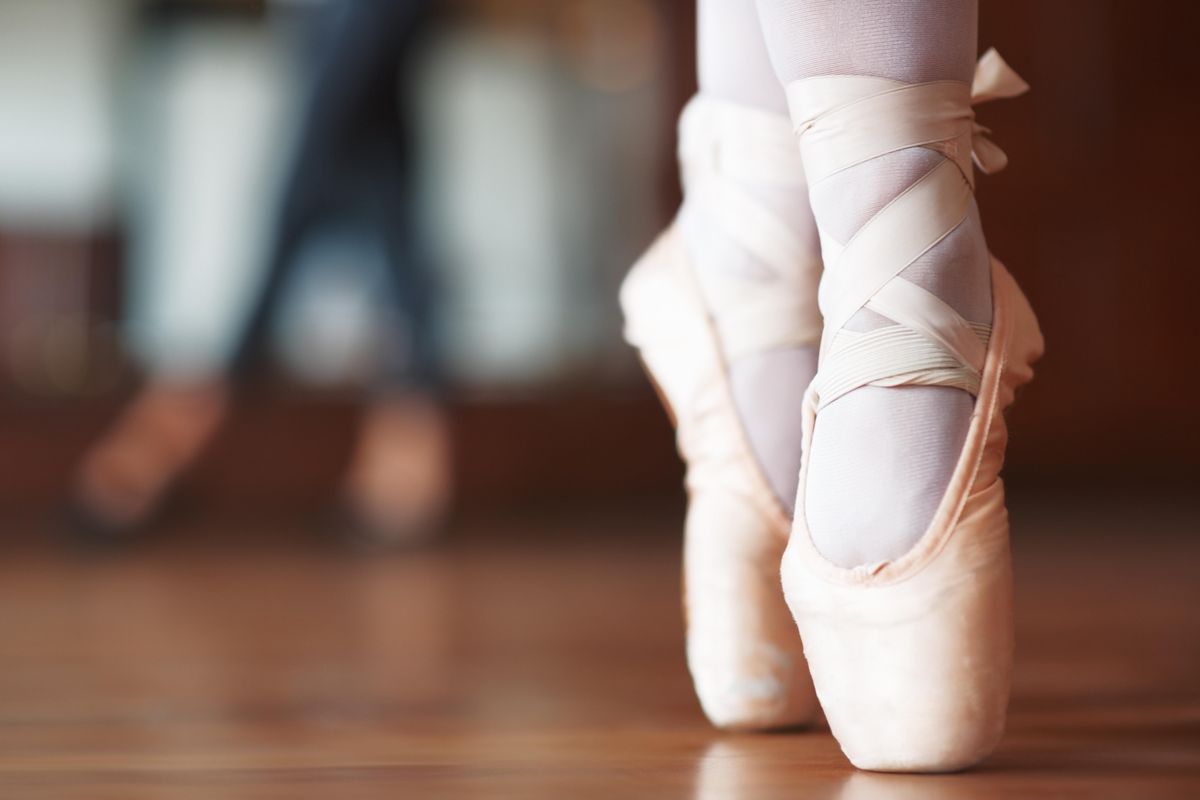A dancer's feet shown from the ankle down, in releve on pointe. The background is out of focus, but seems to show a studio with a window and another dancer