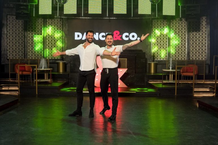Maks and Val Chmerkovskiy, both white men in their 30s, pose with their arms wide and smile in their studio, with a large Dance & Co logo behind them