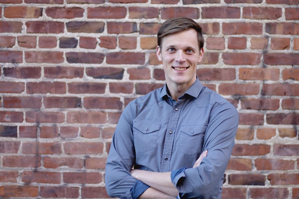 Ben Henry-Moreland, a young white man, wears a blue button-down shirt. He crosses his arms and smiles against a brick wall backdrop.