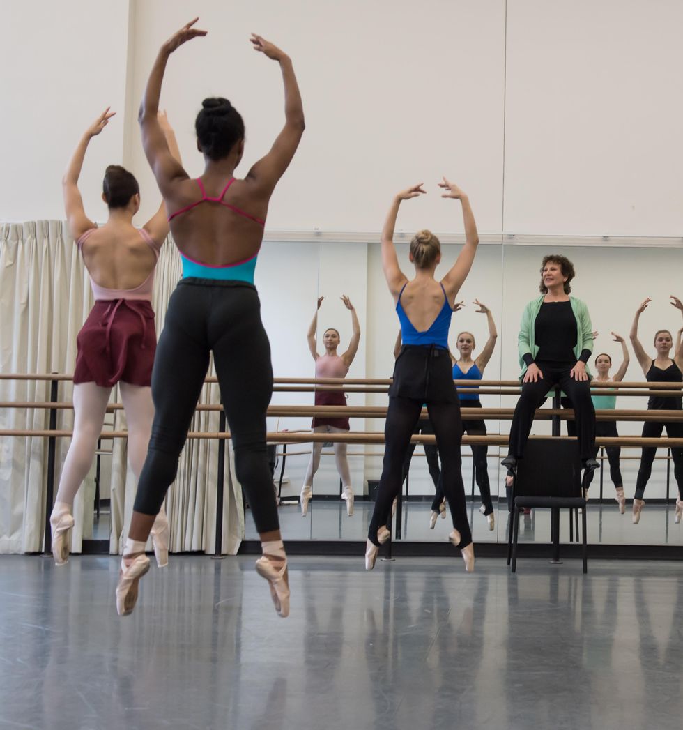 Genn sits on the barre at the front of the studio smiling as dancers jump in second position, arms in fifth