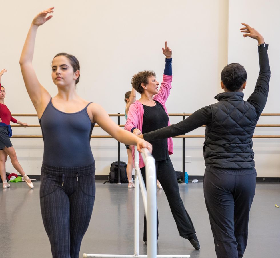 Genn stands at the barre, wearing black pants, a black shirt and a pink sweater, and tendues front, arm raised in fifth position. Dancers at barres surrounding her do the same