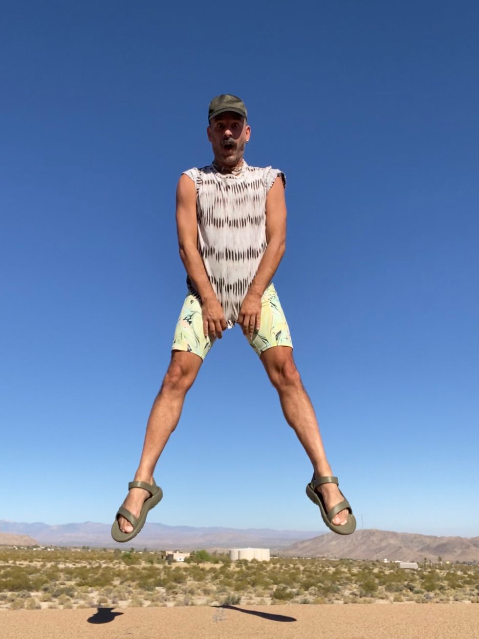 Heffington jumps into the air in his backyard in the desert, making an animated surprised face. He wears sandals, yellow shorts and a black and grey patterned shirt, and a black cap.