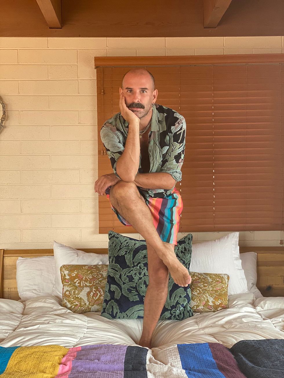 Heffington squats on his bed, wearing colorful striped shorts and a blue and green button down. He has colorful blankets and pillows on his bed