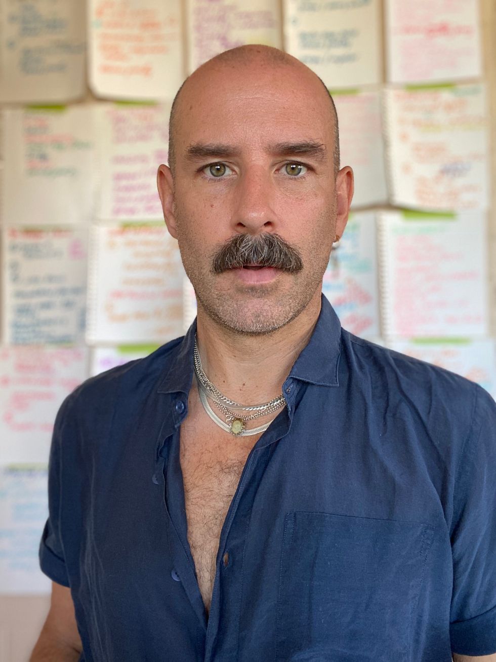 A close-up portrait of Heffington, wearing a dark blue button down and staring intently into the camera. Behind him, a wall plastered with papers that have notes for his virtual classes