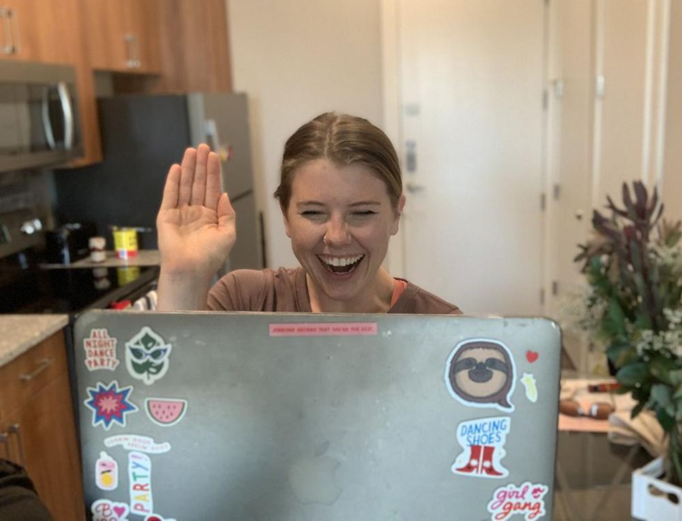 Shannon Oleson sits at her kitchen counter, waving at her computer screen and smiling. Her laptop is covered in colorful stickers, and her kitchen can be seen behind her.