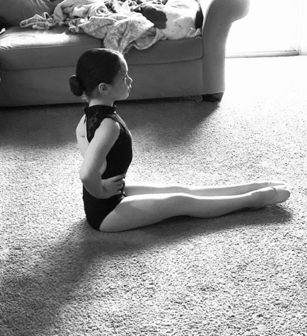 A young girl takes dance class in her living room, wearing a leotard and sitting up very straight with her legs outstretched in front of her