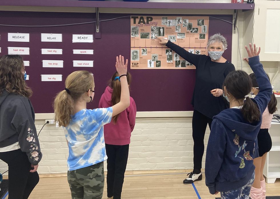 Thelma Goldberg, a older white woman with short grey hair, stands in front of a bulletin board, pointing to an orange timeline that says "tap into history." Young students stand around her, raising their hands.