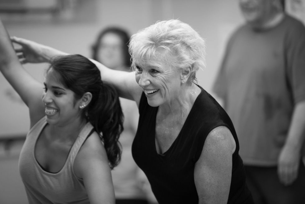 Thelna Goldberg adjusts a student's arm positioning from behind, smiling and looking forward as if there is a mirror there. The student, an adult woman, seems to be laughing.